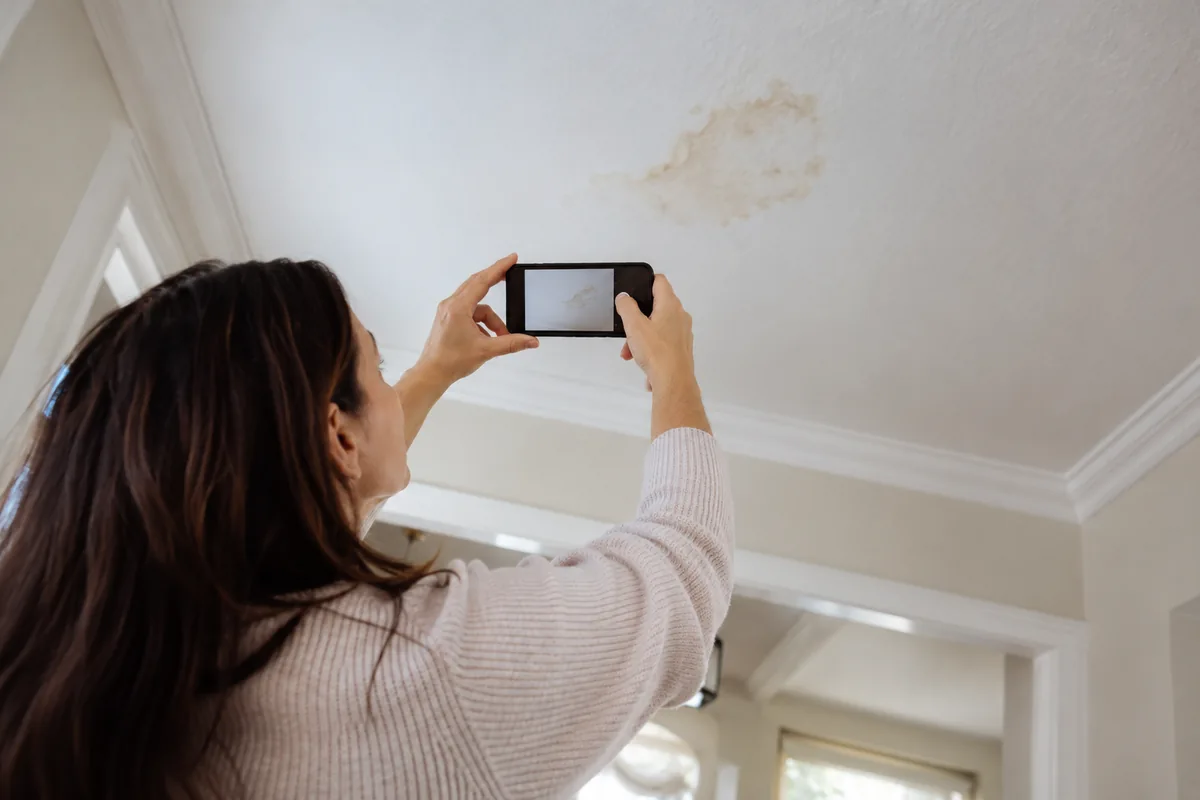 Homeowner photographing a ceiling stain from a safe floor-level position